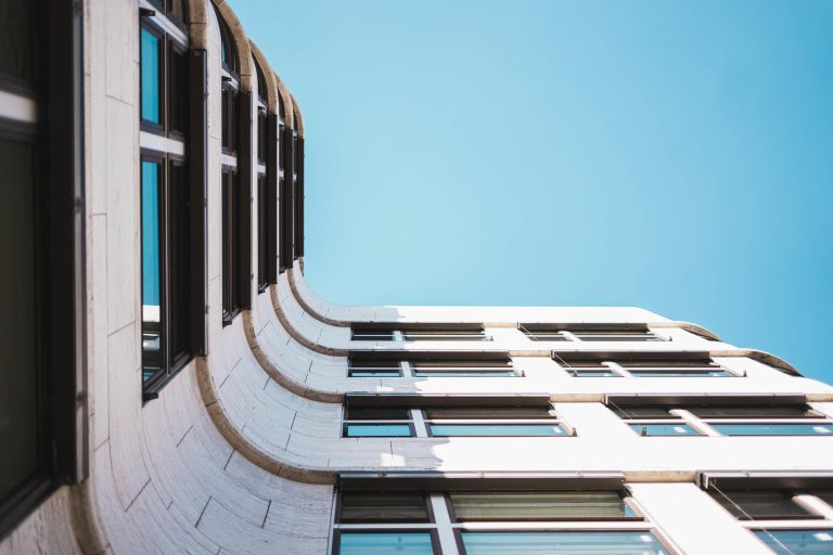 Looking up at a modern white building with a curved facade and dark window frames against a clear blue sky.