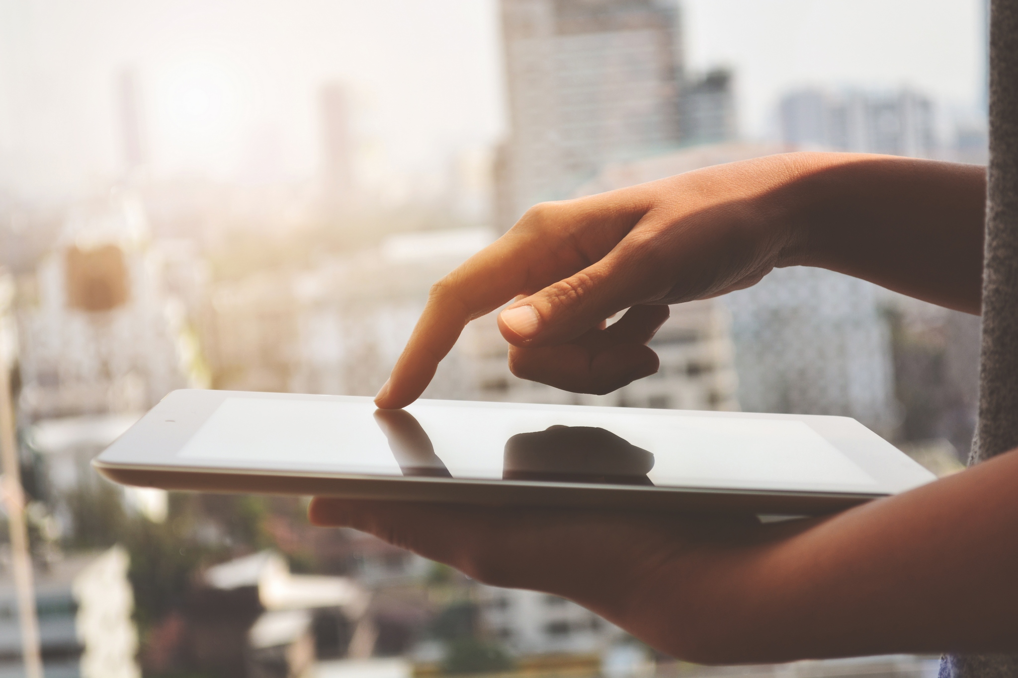Hands holding a tablet displaying a real estate app with property listings, in front of a warmly lit modern building.