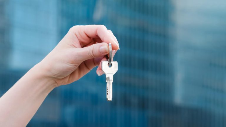 Hand holding a key in front of a modern blue glass building, representing One Eleven's home handover in Larnaca.